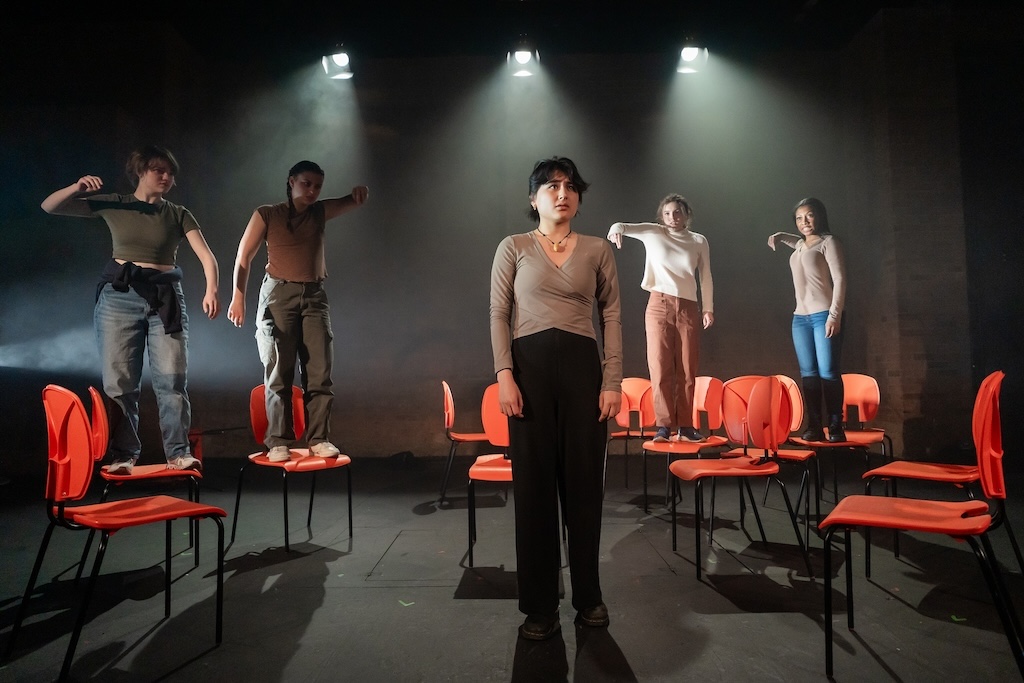 Action shot of the spring 2024 Shakespeare in Performance at RADA final presentation of Julius Caesar. An actor stands in the middle of the stage looking concerned as four actors standing on bright orange chairs behind her, pointing in shame.